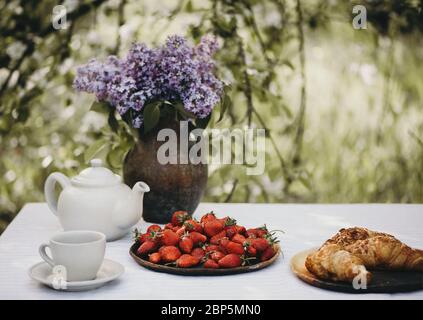 Tasse Tee, Croissants und frische Erdbeeren mit lila Blumen auf dem Tisch im Garten serviert. Stockfoto