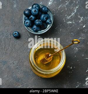 Gesunde Lebensmittel Zutaten - Heidelbeeren und Honig. Glasgefäß mit Honig und Dipper, frische Heidelbeeren in kleiner Schüssel auf dunklem Hintergrund. Draufsicht oder flaches Lay. Quadratisches Zuschneiden. Kopierbereich Stockfoto