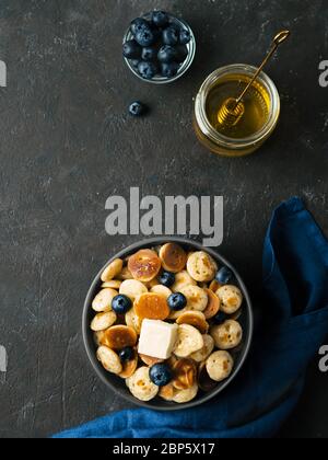 Trendige Küche: Pfannkuchen, Müsli, Butter und Heidelbeeren. Haufen Mini-Cereal-Pfannkuchen in Boule auf dunklem Hintergrund. Kopieren Sie den rechten Platz für Text. Draufsicht oder flaches Lay. Vertikal. Stockfoto