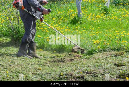 Ein Bauer mäht mit einem Trimmer Gras mit Löwenzahn in einem Gartengrundstück. Nahaufnahme eines Arbeiters in Spezialkleidung mit einem Gasmäher in der Hand. Stockfoto