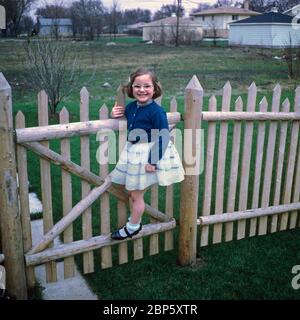 Ein glückliches junges Mädchen in ihrem Partykleid schwingt aus einem Tor in ihrem Garten, USA 1967. Stockfoto