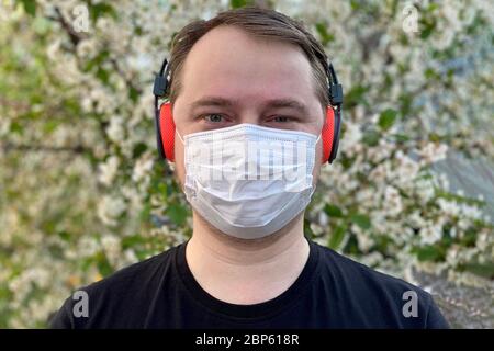 Porträt eines lächelnden jungen Mannes, der die medizinische Grippemaske und die Kopfhörer trägt, auf Blumenhintergrund. Um Coronavirus zu verhindern. Stockfoto