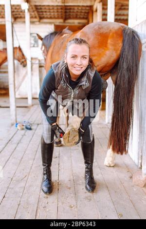 Junge schöne Frau reinigt das Pferd Hufe mit einem speziellen Pinsel vor dem Reiten. Reiten, Tierpflege, tierärztliche Konzept Stockfoto