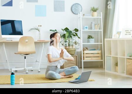 Ganzkörperportrait der zeitgenössischen jungen Frau beim Yoga, während sie in Lotusstellung zu Hause sitzt, kopieren Raum Stockfoto