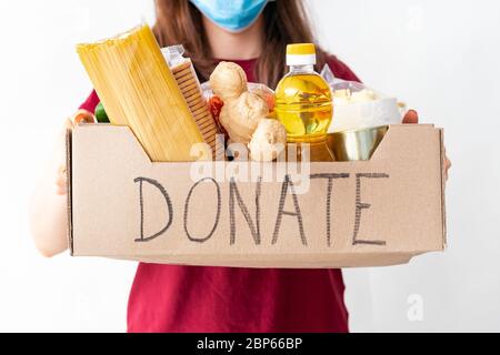 Spendenbox mit Essen. Frau in schützenden Gesichtsmaske hält Spenden Box voller Nahrung auf weißem Hintergrund. Freiwillige machen Lebensmittel Lieferung während der covid Stockfoto