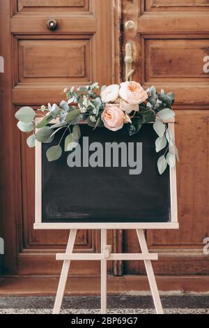 Leere Hochzeit Tafel Schild Mockup Szene. Holzstaffelei mit Willkommenstafel. Florale Girlande aus grünen Eukalyptuszweigen und englischen Rosen Blüten. Ru Stockfoto