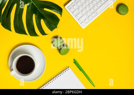 Lebendiger Arbeitsplatz mit Tastatur, Notebook, Stift, Tasse Kaffee, grünen Macarons und Monstera-Blatt auf gelbem Hintergrund. Home Office, freiberufliche Arbeit oder blo Stockfoto