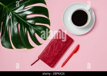 Weiblicher Arbeitsplatz mit rotem Notizbuch und Stift, weiße Tasse schwarzen Kaffee und grünes Monstera-Blatt auf rosa Hintergrund. Home Office, Tagebuch oder Planung conce Stockfoto