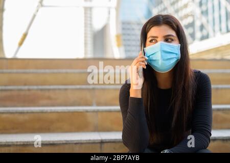 Junge indische Frau mit Maske am Telefon und sitzt an der Treppe in der Stadt Stockfoto