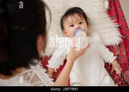 Baby Mädchen trinken Milch aus der Flasche, die Mama gibt ihr und Blick auf die Kamera Stockfoto