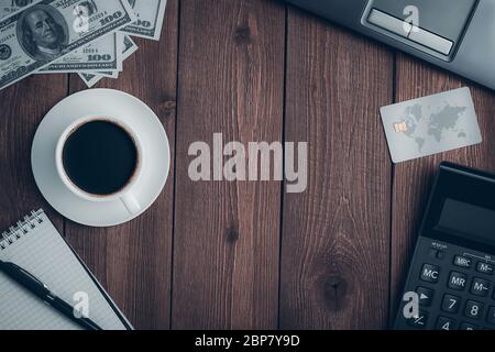 Draufsicht auf einem rustikalen Holztisch mit einer Tasse Kaffee, einer Kreditkarte, einem Taschenrechner, einem Notizbuch mit Stift, Dollarscheinen und einem Laptop mit Kopierplatz. Stockfoto
