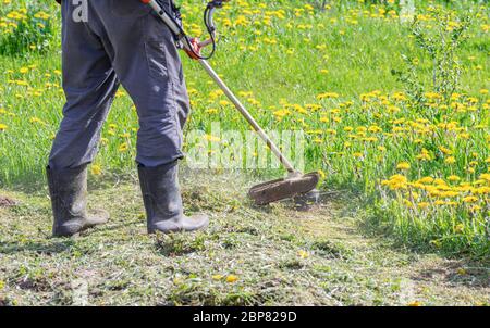 Ein Bauer mäht mit einem Trimmer Gras mit Löwenzahn in einem Gartengrundstück. Nahaufnahme eines Arbeiters in Spezialkleidung mit einem Gasmäher in der Hand. Stockfoto