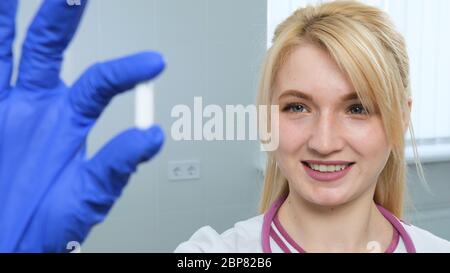 Nahaufnahme von kaukasischen jungen blonden weiblichen Sandiktäter in weißem Fell, Stethoskop hält eine weiße Pille mit Fingern in blauen Handschuhen im Krankenhaus oder Stockfoto
