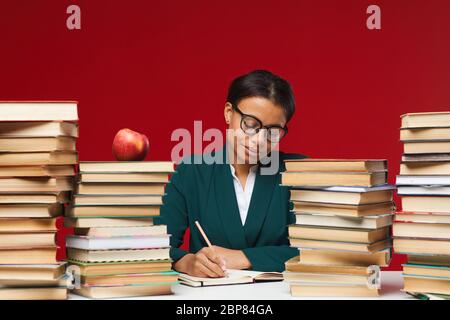 Vorderansicht Porträt der jungen Lehrerin sitzt am Schreibtisch zwischen Stapeln von Büchern vor rotem Hintergrund, Kopie Raum Stockfoto