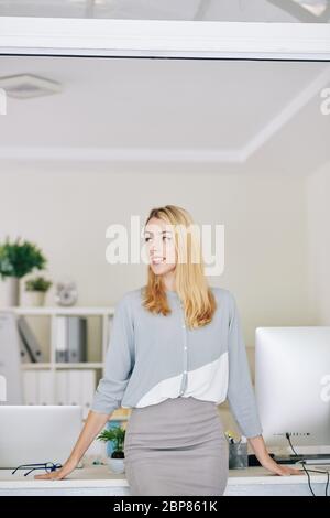 Attraktive junge lächelnde Geschäftsfrau steht am Bürotisch und schaut weg Stockfoto
