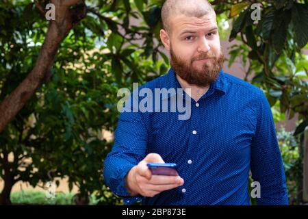 Kahlbärtiger Mann in einem blauen Hemd, der draußen ein Smartphone hält und auf einen zeigt Stockfoto