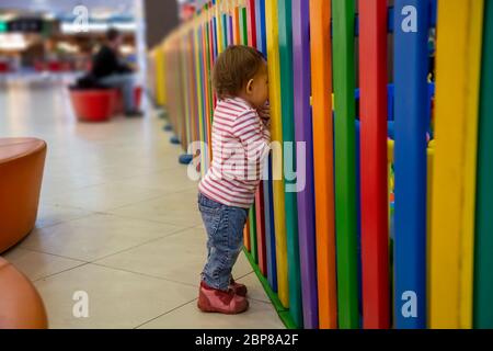 Ein süßes kleines Kleinkind steht am bunten Zaun eines Kinderspielzimmers und schaut durch ihn und will drinnen spielen. Soft Focus. Close-up Stockfoto