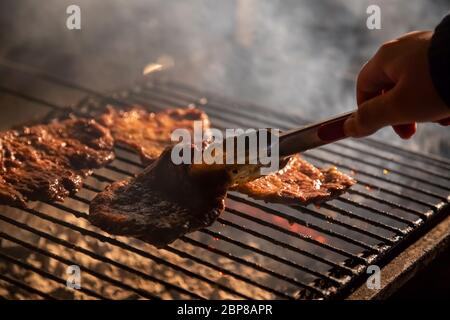 Abends ein Barbecue-Grill, auf dem leckere saftige Steaks über offenem Feuer gegrillt werden, eine Hand hält die Zange und dreht das Fleisch um. Nahaufnahme, weich Stockfoto