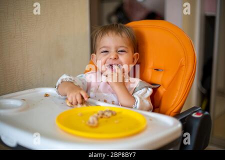 Niedliches Baby in einem orangefarbenen Kindersitz schaut die Kamera lachend an und zeigt Hand auf Zähne. Die Zähne sind gewachsen. Krümel und eine orangefarbene Platte mit einem Kuchen auf der Stockfoto