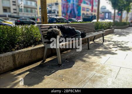Obdachloser Bettler schmutziger Mann schläft auf einer Bank unter einem Baum auf der Straße, im Hintergrund die Stadt, Verkehr, Einkaufszentrum und bunten Häusern in Stockfoto