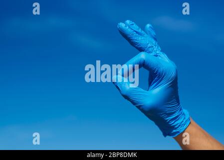 Nahaufnahme der Hand eines Mannes, mit blauen OP-Handschuhen, das OK-Zeichen gegen den blauen Himmel, mit etwas Leerzeichen auf der linken Seite Stockfoto