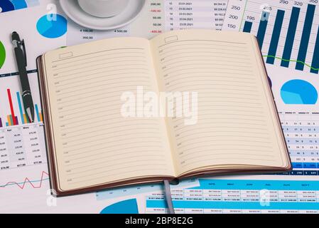 Die Arbeitskomposition. Ein Büro-Desktop mit einer leeren Tagebuchseite. Statistische Grafiken, Tabellen mit Zahlen und Geld, eine Tasse Kaffee und einen Stift. Stockfoto