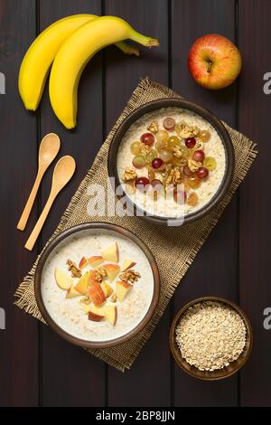 Haferbrei Haferbrei mit Trauben, Äpfeln und Walnüssen in rustikalen Schalen, frischen Apfel, Bananen, Holzlöffel und rohen Haferflocken an der Seite, fotografiert Overhe Stockfoto