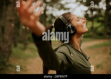 Hübsche junge Frau mit Kopfhörer, die ihre Arme im Wald vorpreading, weil sie draußen trainiert Stockfoto