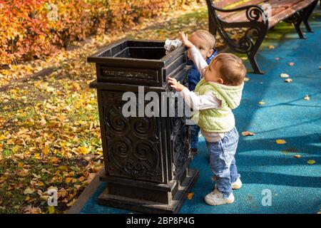 Zukunft Ökologie Konzept. Zwei kleine süße Babys stehen in der Nähe der schwarzen Straße Container im Park und werfen Müll dorthin. Ein weiteres Kind kommt Stockfoto