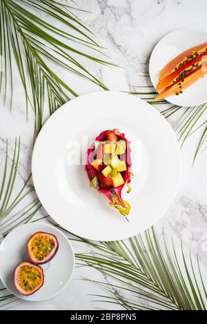 Flache Plattenlage mit gemischten Früchten und geschnittenen Früchten. Tropischer Obstsalat in halber Drachenfrucht. Drachenfrucht, Maracuja, Papaya, Palmblätter Stockfoto