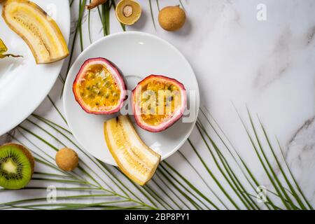 Geschnittene Passionsfrucht, Londan, Banane auf weißem Gericht. Exotische Früchte und tropische Blätter auf weißem Marmorhintergrund mit Kopierraum für Ihren Text. Lebensmittel, Stockfoto