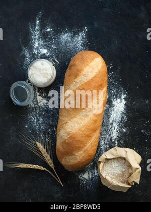 Britisches Weißblüher oder europäischer Sauerteig Baton-Brot auf schwarzem Hintergrund. Frisches Brot und Glas mit Sauerteig Starter, floer in Papiertüte und Ohren. Draufsicht. Kopierbereich. Vertikal. Stockfoto