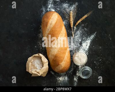Britisches Weißblüher oder europäischer Sauerteig Baton-Brot auf schwarzem Hintergrund. Frisches Brot und Glas mit Sauerteig Starter, floer in Papiertüte und Ohren. Draufsicht. Kopierbereich Stockfoto