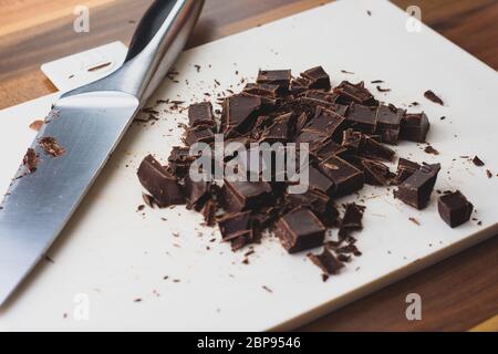 Schokolade, die auf einem weißen Schneidebrett in grobe Würfel geschnitten wurde. Das Messer, das zum Schneiden der Schokolade verwendet wird, ist ebenfalls im Rahmen Stockfoto