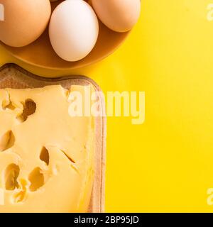 Stück Käse auf einem Brett mit Eiern auf gelbem Hintergrund mit Kopierraum. Stockfoto