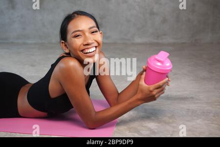 Glückliche gesunde lächelnde Frau hält Protein Shake entspannend auf der rosa Yoga-Matte im Fitnessstudio. Frau in Sportkleidung posiert im Fitnessstudio Stockfoto