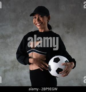 Porträt von schönen jungen lächelnden Frau in Sportkleidung hält Fußball isoliert über Beton Hintergrund Stockfoto