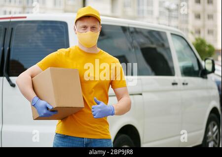 Lieferung Mann Mitarbeiter in gelben Uniform Kappe, T-Shirt, Gesichtsmaske, Handschuhe hält eine Kartonverpackung steht nar Lieferwagen . Sicherheit Lieferung Quaran Stockfoto