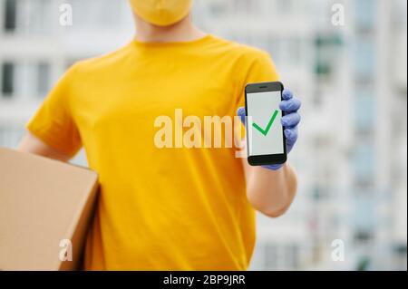 Lieferung Mann Mitarbeiter in gelben Uniform Mütze, T-Shirt, Gesichtsmaske und Handschuhe hält eine Kartonverpackung zeigt komplette Zeichen auf dem Telefon. Sicherheitskost Stockfoto