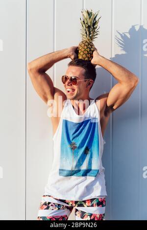 Gerne trendy Mann in Sonnenbrille hält eine Ananas auf dem Kopf, und das Lächeln, das Tragen von stilvollen Leibchen und Hosen. Im Freien. Stockfoto