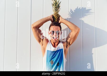 Freude schöner Mann in Sonnenbrille hält eine Ananas auf dem Kopf, in die Kamera schauen und lächeln, das Tragen von stilvollen singlet. Im Freien. Stockfoto