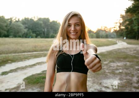 Schöne lächelnde Mädchen der Ohrhörer, ein Daumen hoch, und die Kamera, das Tragen von Sportbekleidung. Im Freien. Stockfoto