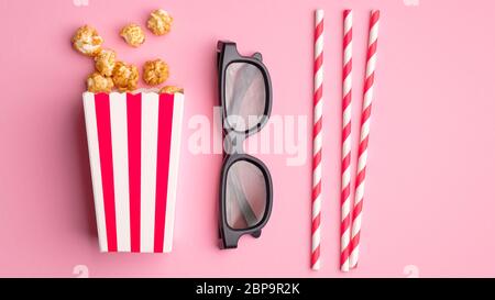 Flache Lay Komposition mit Popcorn in gestreifter Kartonschachtel, 3d-Gläsern und Trinkhalmen auf rosa Hintergrund. Film im Kino-Konzept ansehen. Stockfoto