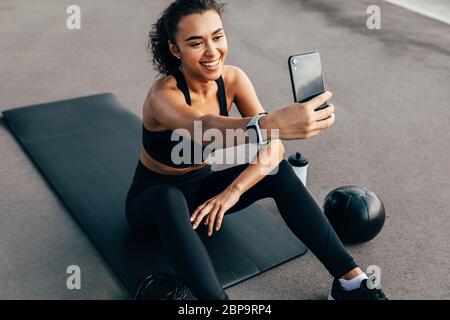 Sportlerin sitzt auf einer Matte im Freien und nimmt Selfie für soziale Medien auf. Lächelnde Frau, die abends während des Trainings eine Pause einbricht. Stockfoto