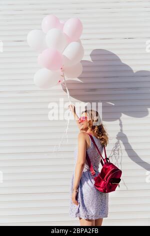 Rückansicht des glücklichen Mädchen in der Sonnenbrille, Luftballons, das gegenüber der weißen Wand, im Freien. Im Sommer Kleid gekleidet, mit Rucksack. Stockfoto