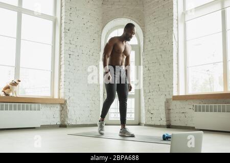 Junger afroamerikanischer Mann trainiert zu Hause während der Quarantäne des Coronavirus Ausbruch, doinc Übungen der Fitness, Aerobic. Sportivfähigkeit während der Isolierung. Wellness, Bewegungskonzept. Stockfoto