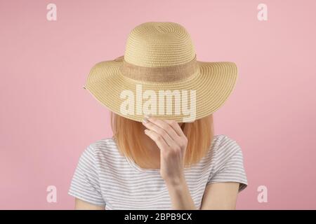 Rothaarmädchen in grauem T-Shirt versteckt sich hinter einem Strohhut, während sie ihn mit ihren Händen über ihr Gesicht zieht. Frontansicht Oberkörper rosa Stockfoto