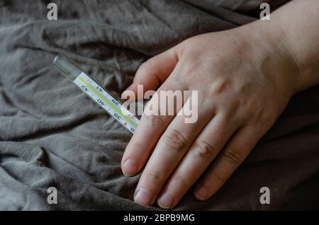 Weibliche Hand liegt auf einer Decke. In der Hand befindet sich ein Quecksilberthermometer. Das Konzept ist Bettruhe, Behandlung, Temperaturmessung. Aufnahmen auf Augenhöhe. Stockfoto
