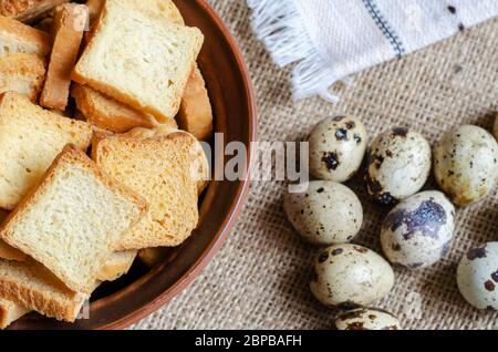 Stillleben mit Croutons und rohen Wachteleiern auf Burlap. Eine Tonschüssel mit kleinen quadratischen Semmelbröseln und einer Gruppe Wachteleier. Einfache gesunde Lebensmittel. Vie Stockfoto