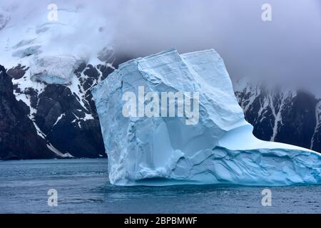 Ein großer blauer Eisberg, der wie ein Hausschuh geformt ist, schwimmt langsam von einer nebelbedeckten Elefanteninsel auf dem Kontinent der Antarktis. Stockfoto